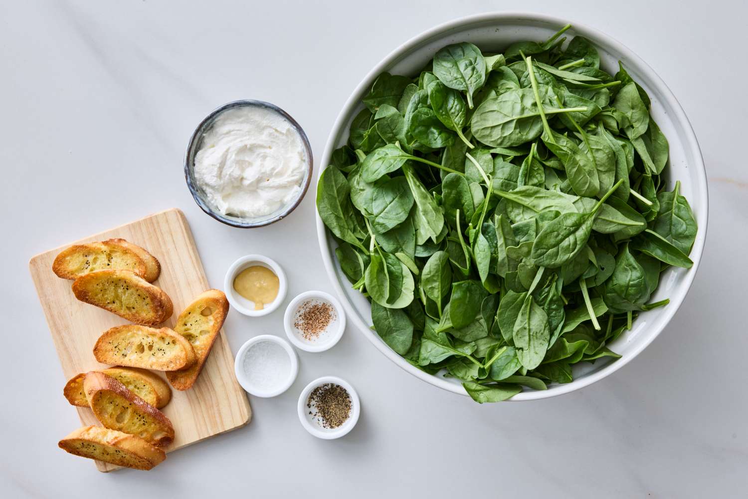 A bowl of fresh spinach leaves a small dish of cream and toasts arranged on a white surface