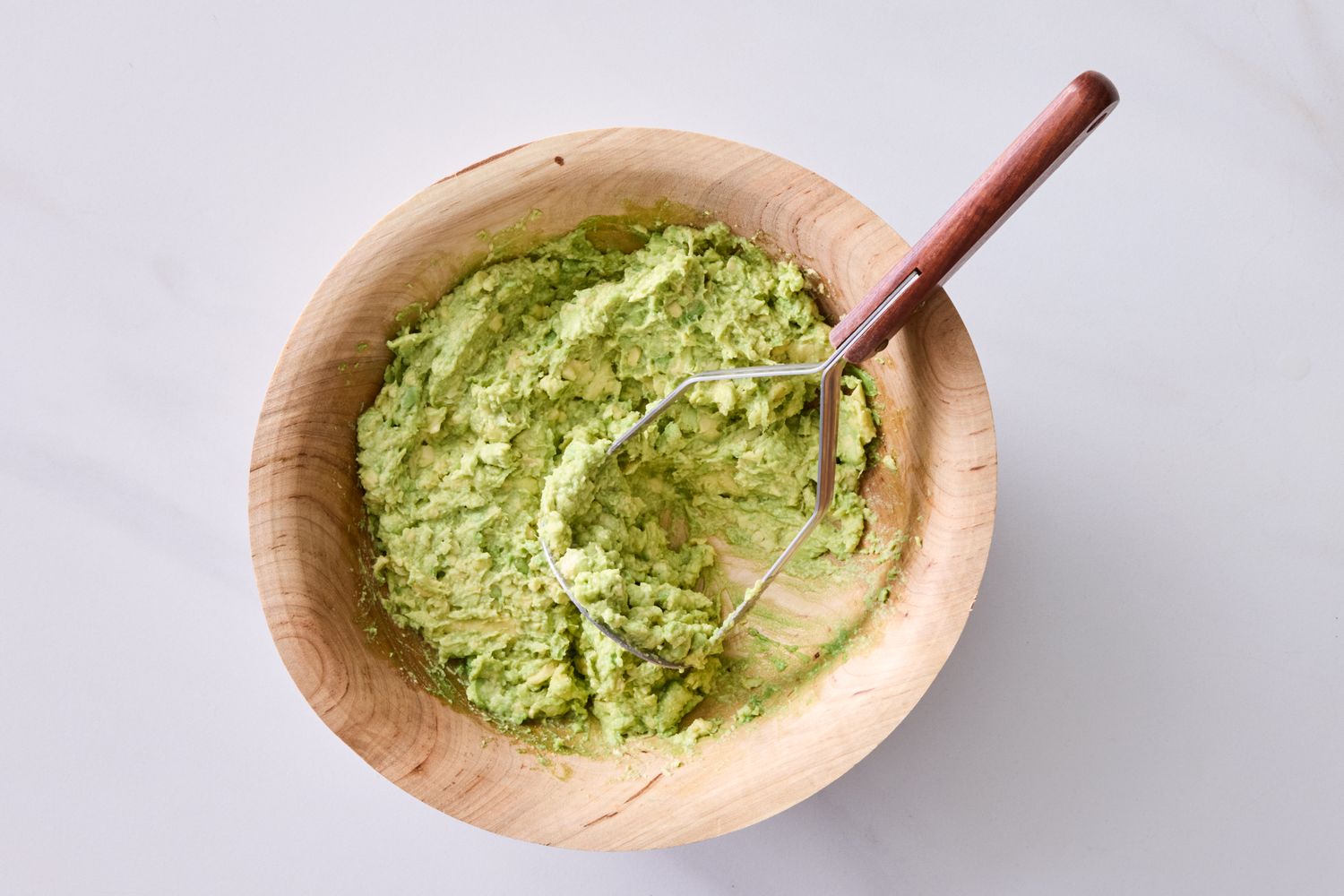 A wooden bowl containing mashed guacamole with a masher