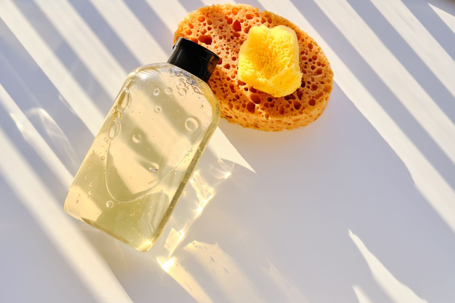 A bottle of clear liquid soap and a sponge resting on a white surface with light streaming through blinds