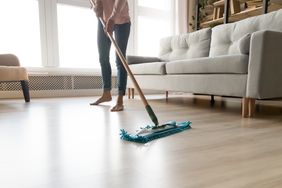 Person mopping a hardwood floor in a living room with a couch and a window in the background