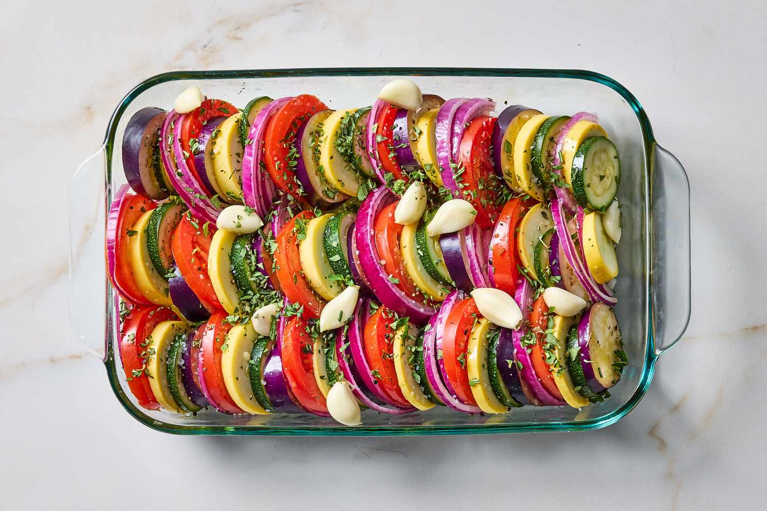 Layered vegetable bake in a clear glass dish featuring slices of zucchini tomato squash and onion with garlic and herbs