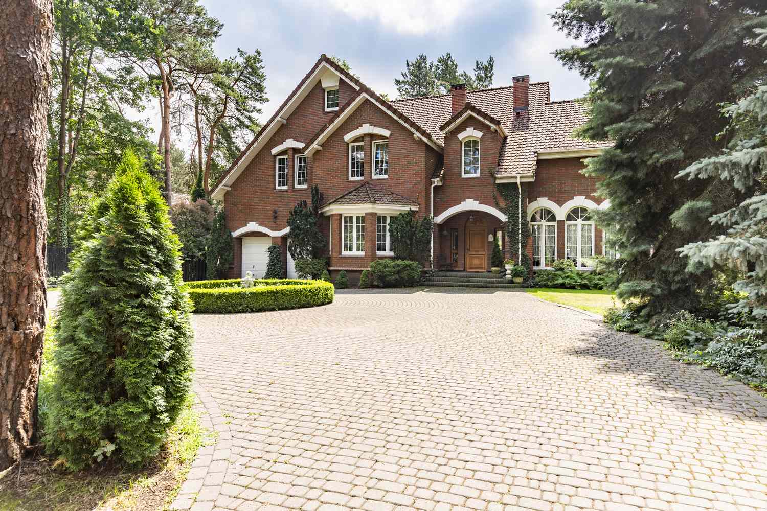 A large brick house with a circular driveway surrounded by trees and shrubs in a forested area