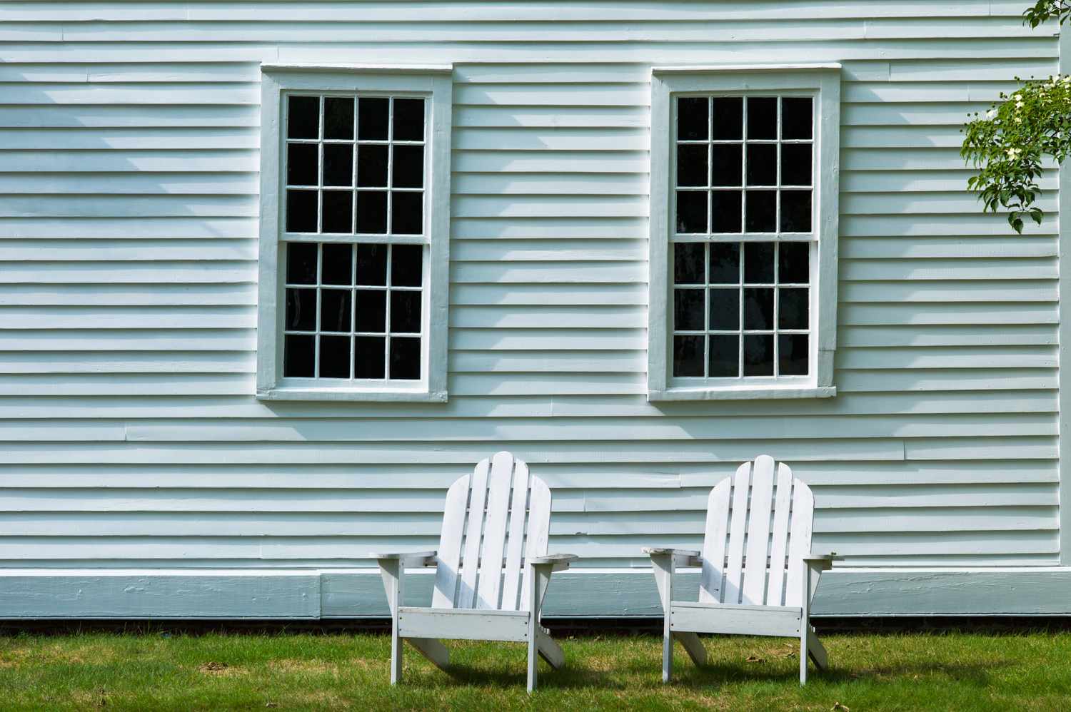 Two Adirondack chairs on the lawn outside a white clapboard house with two windows