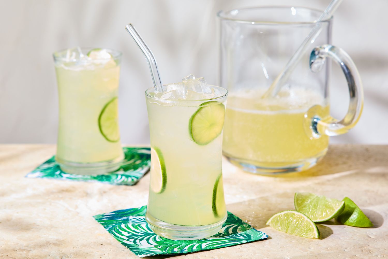 Two glasses of lime squash drink with ice next to a pitcher and lime slices
