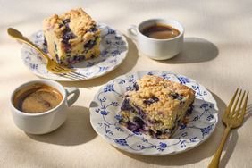 Two servings of blueberry crumb cake on patterned plates with cups of coffee and golden forks