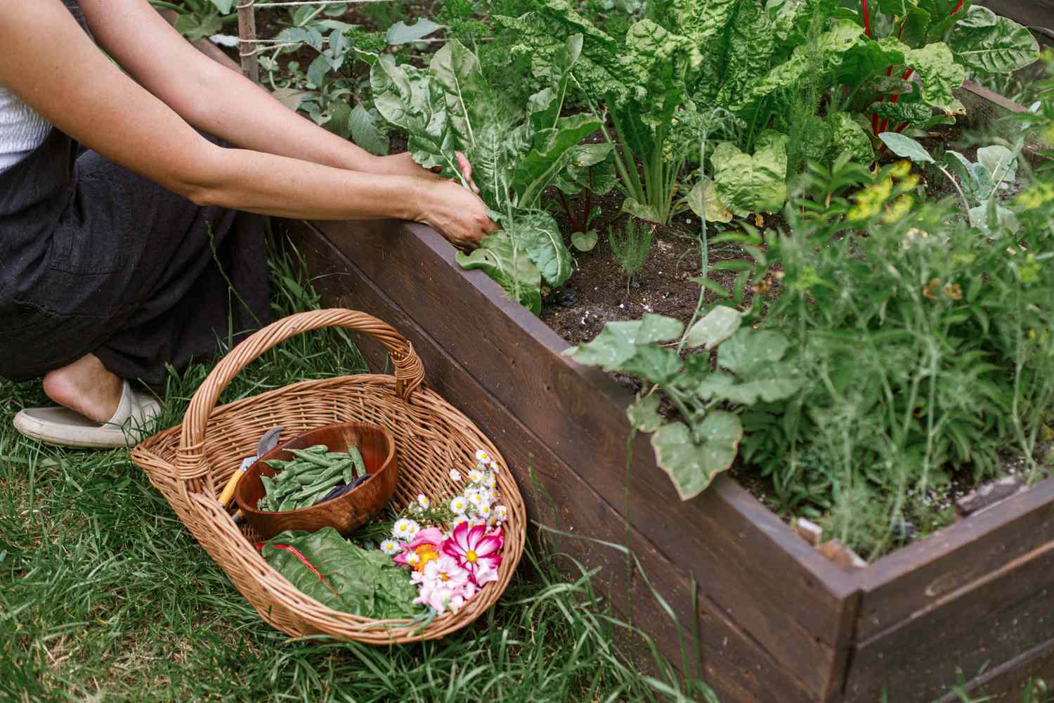Person tending to a garden bed with a basket of harvested produce and flowers placed nearby