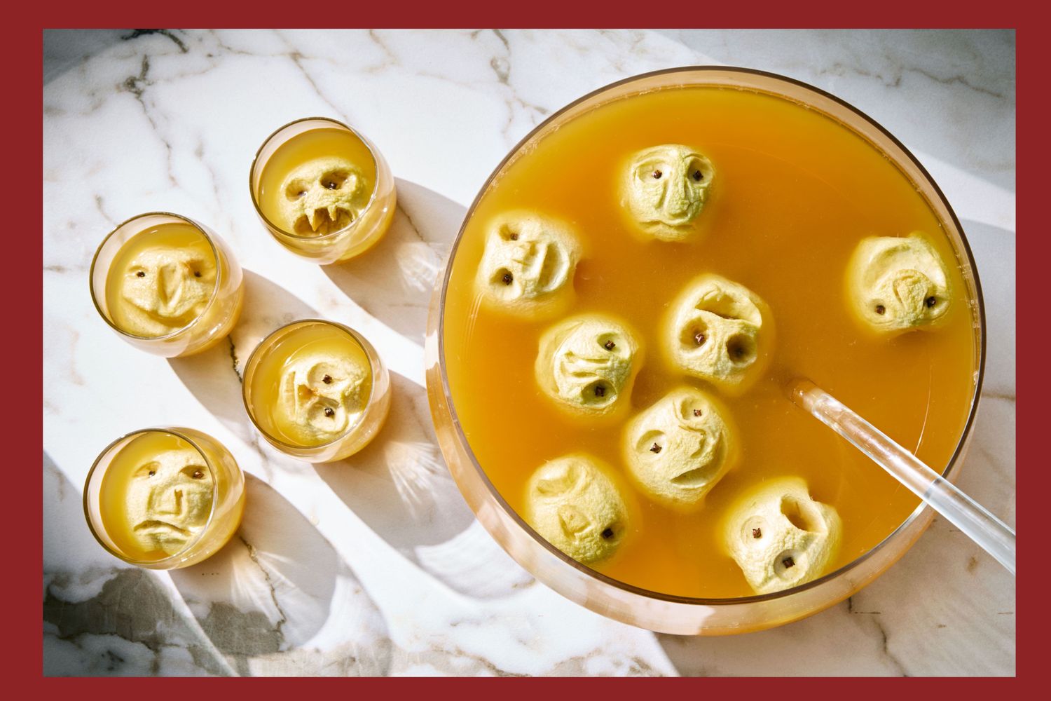 A bowl of orange punch with floating shrunken head decorations and five matching glasses on a marble surface themed for Halloween