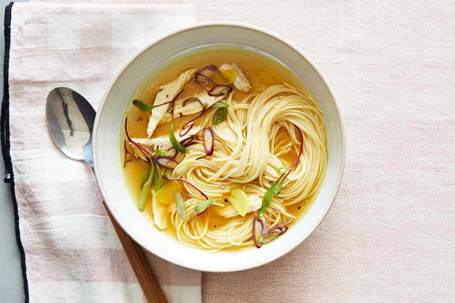 A bowl of noodle soup served with slices of ginger and garnish placed on a napkin with a spoon beside it