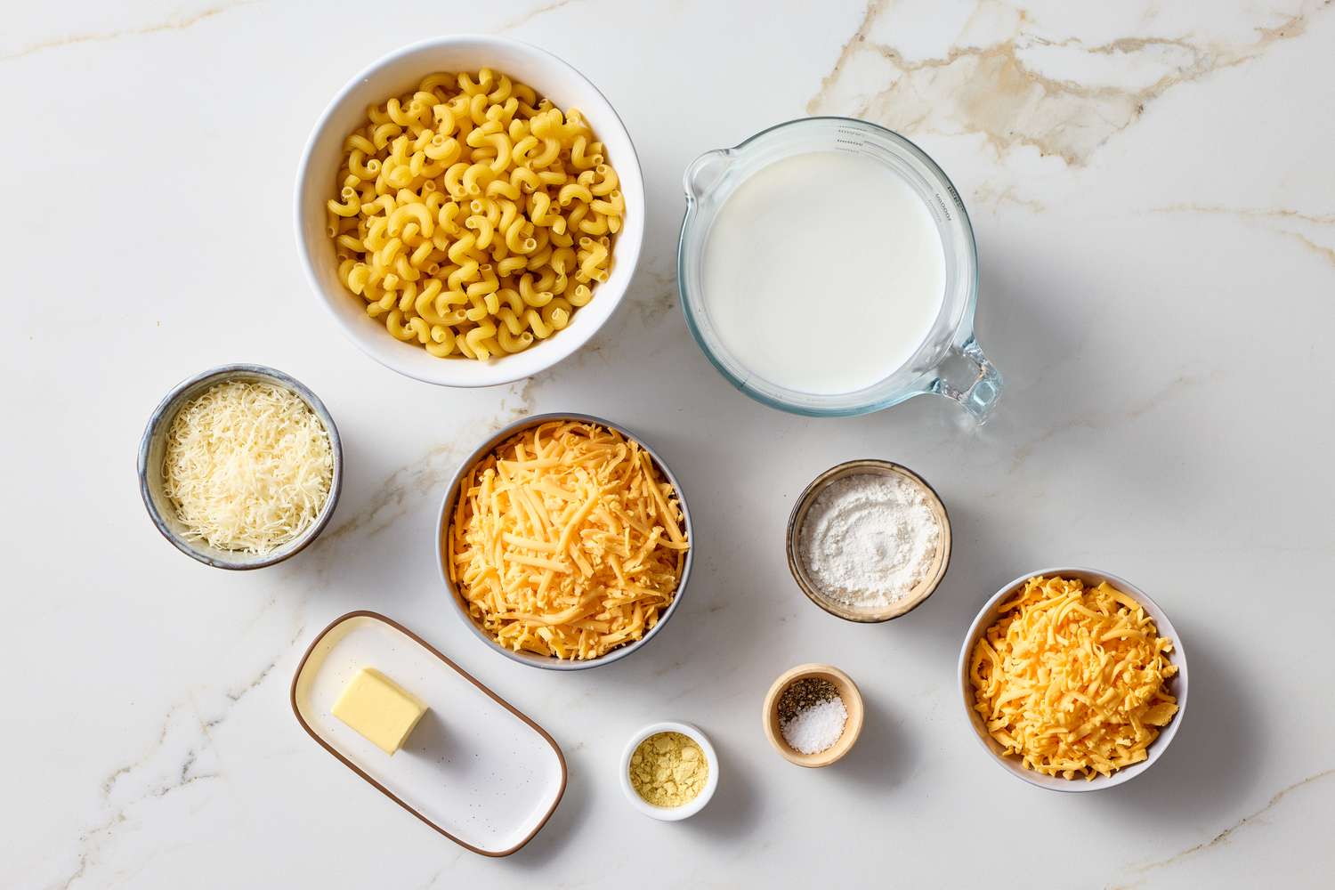 Ingredients for mac and cheese including pasta grated cheese milk butter and spices arranged on a marble surface