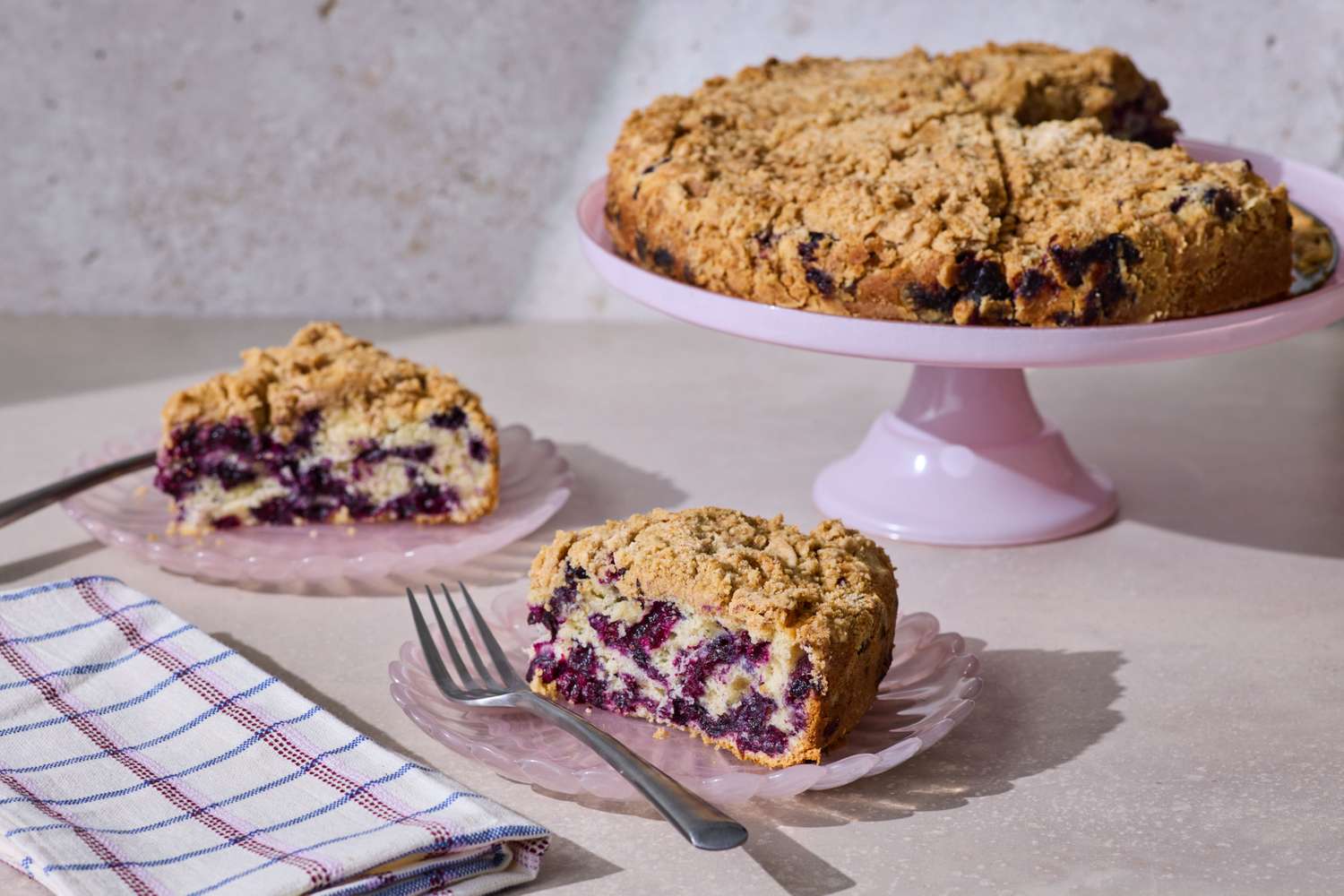 A blueberry buckle cake on a pink stand with two plated slices and utensils nearby