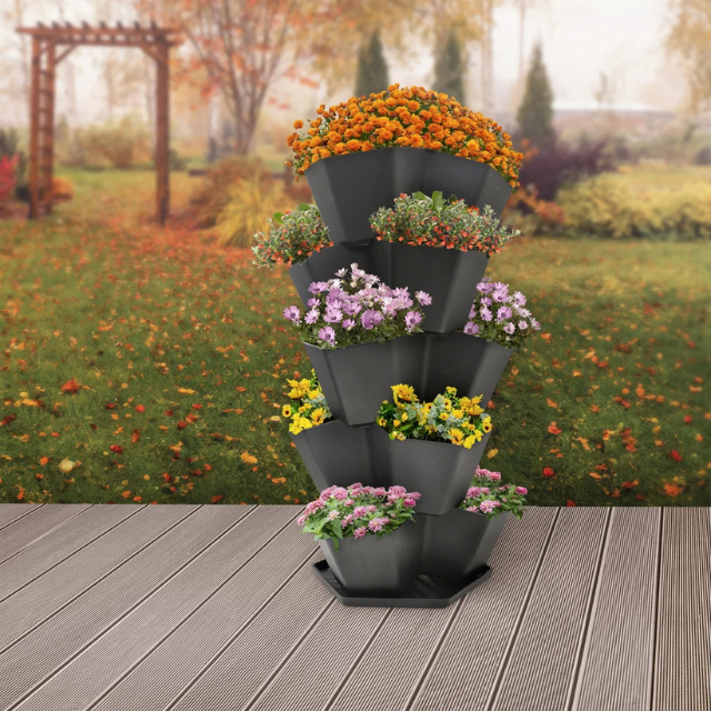 A multitiered flower planter holding various flowers on a patio with a garden and trees in the background during autumn