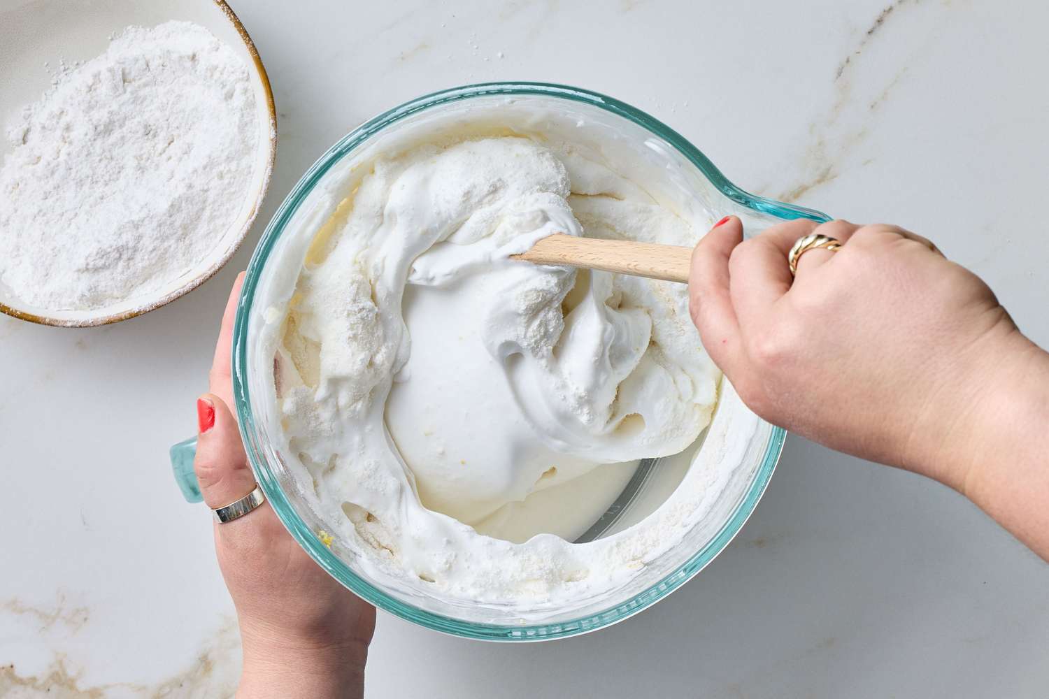 Hand stirring whipped batter in a glass mixing bowl with a spatula, a bowl of flour nearby