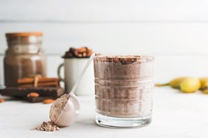 A glass of a smoothie or protein shake with a scoop of powder with additional ingredients blurred in the background