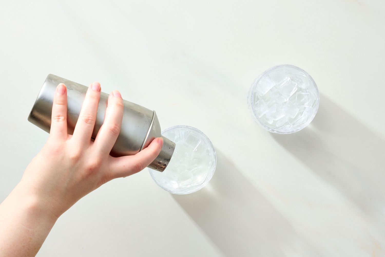 A hand pouring liquid from a shaker into a glass filled with ice with another glass of ice nearby