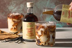 An iced beverage being prepared with vanilla bean syrup with syrup bottles and glasses on a table