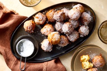 A platter of apple fritters dusted with powdered sugar with a small container of sugar and additional fritters on a side plate