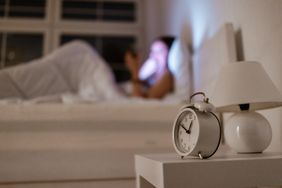 An alarm clock on a bedside table with a blurred figure lying in bed in the background