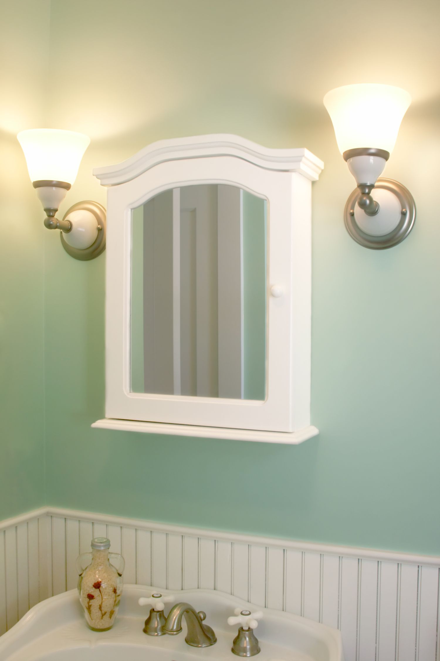 A white bathroom cabinet with a mirror mounted on a green wall flanked by two sconces
