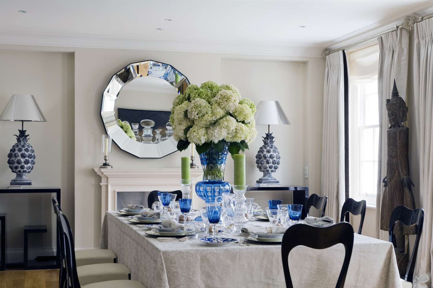 A formal dining table with a centerpiece of flowers and table settings a round mirror above a mantel in the background