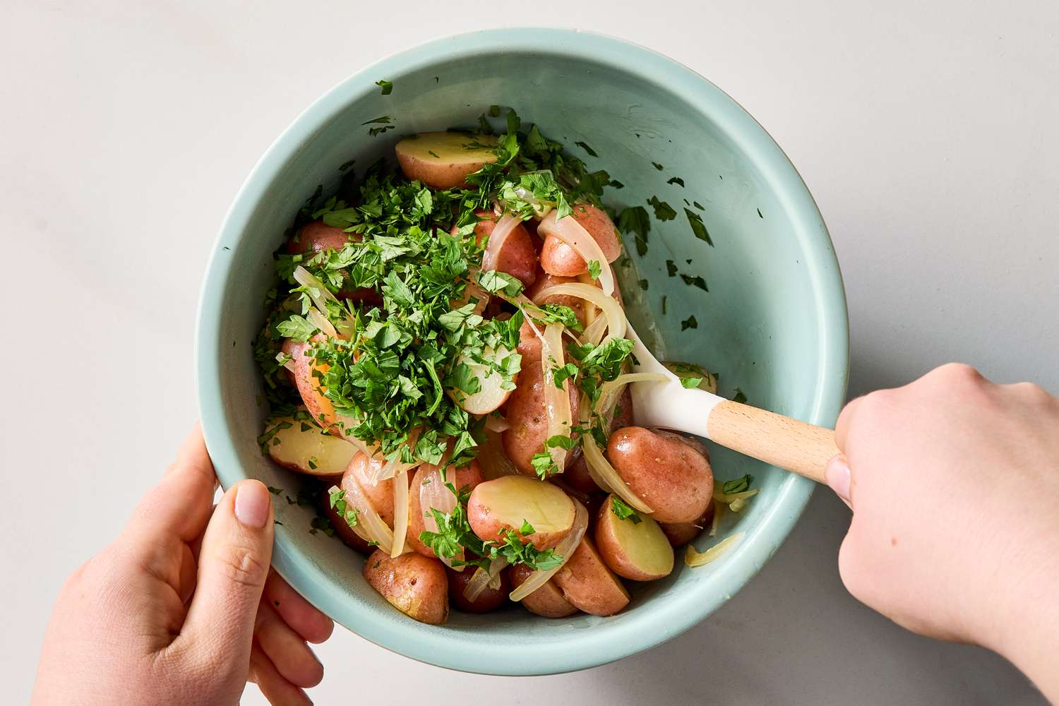 A bowl containing cooked potatoes mixed with herbs held by two hands using a spoon