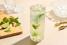 A mojito cocktail in a glass, mint leaves and lime pieces on a wooden board beside