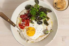 A bowl of savory oatmeal topped with a fried egg greens and cherry tomatoes accompanied by a cup of coffee