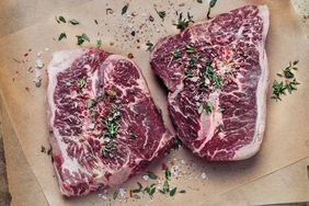 Two raw beef steaks garnished with herbs and seasoning on parchment paper viewed from above