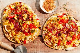 Two grilled pizzas topped with various vegetables and spices, with a pizza cutter and bowl of seasoning on a wooden surface