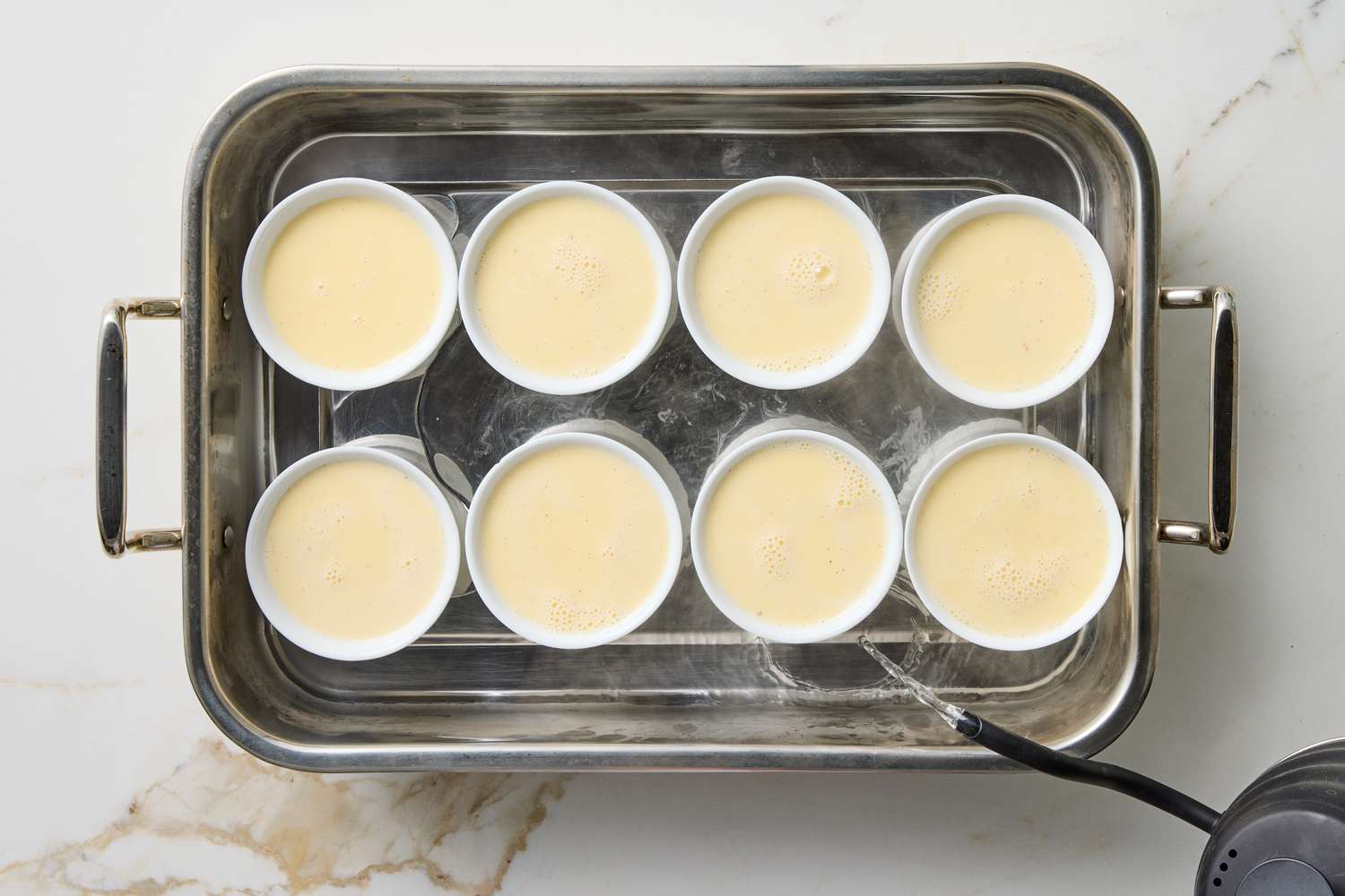 Eight ramekins with custard in a baking dish water being poured for a bainmarie