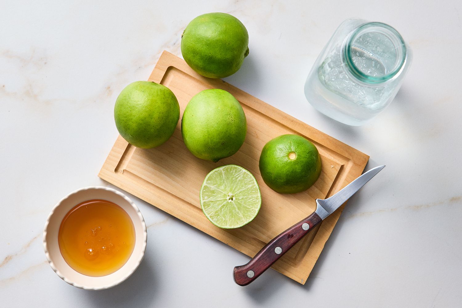 Limes on a cutting board a jar of liquid a knife and a bowl of honey as potential drink ingredients