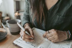 A person writing in a notebook with a pen a mug and desk items in view