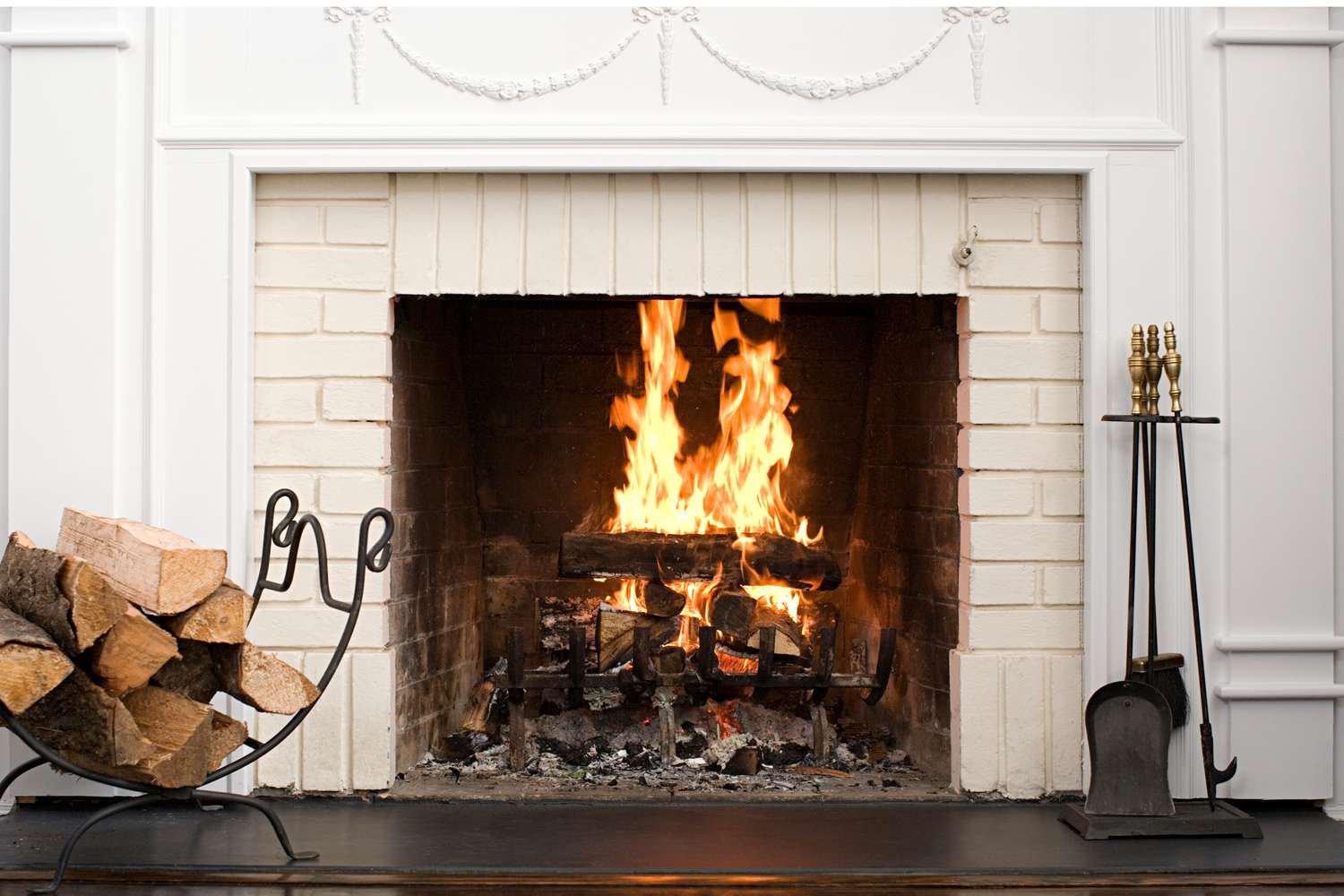 A fireplace with a lit fire wood stacked in a holder nearby and fireplace tools on the side
