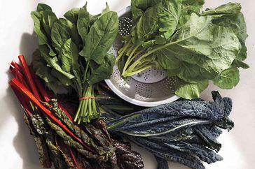 Various fresh leafy greens arranged on a surface including chard spinach kale and collard greens around a colander