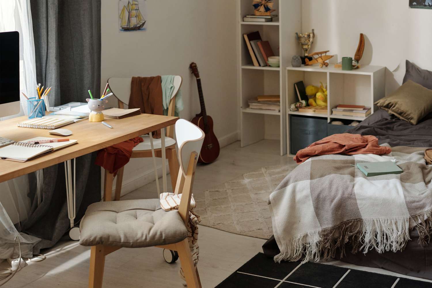 A cluttered bedroom and study area with a desk a chair and a bed visible accompanied by scattered books and other personal items