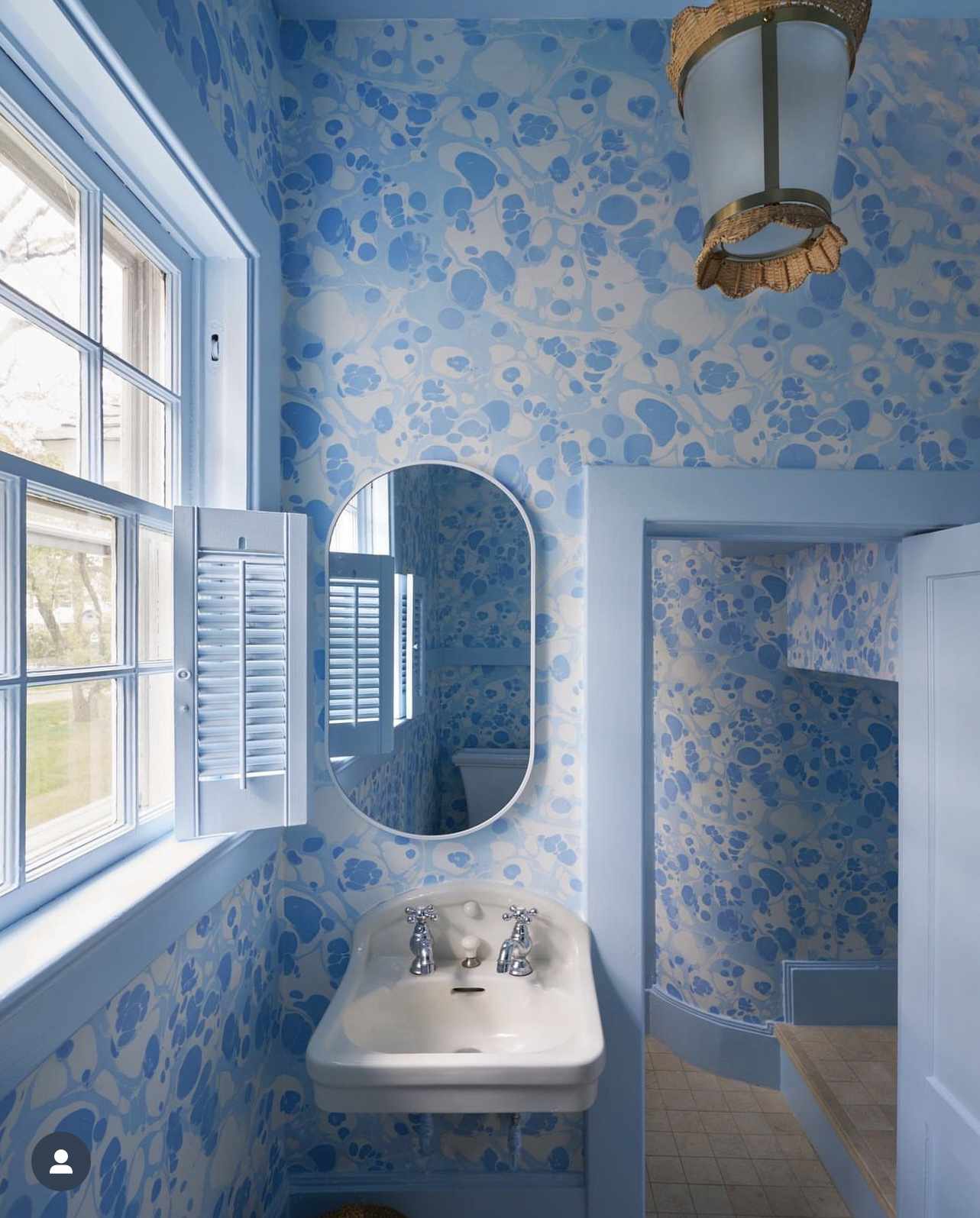 Bathroom with floral wallpaper, an oval mirror, and a small sink under a window with shutters
