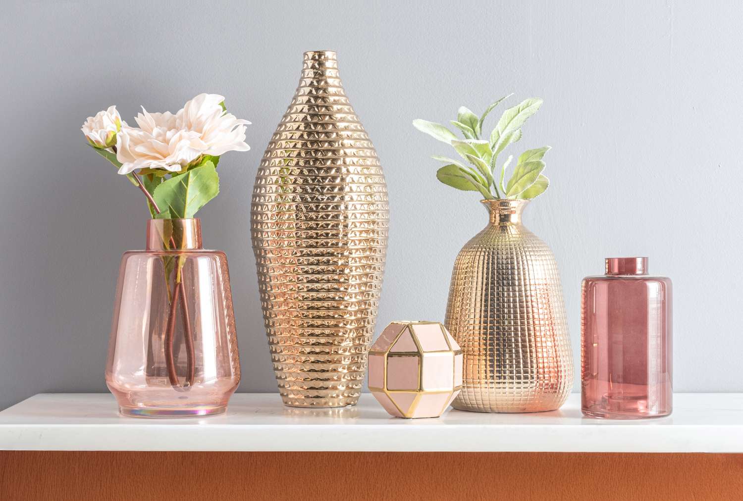 A set of decorative vases and containers some with flowers or greenery arranged on a white shelf