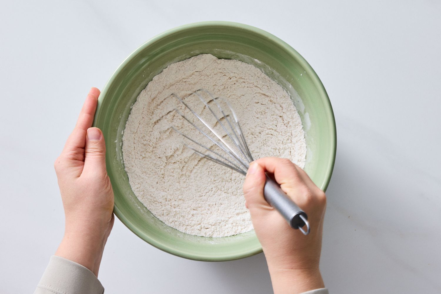 Hands holding a bowl of flour while using a whisk