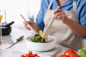 A person preparing a fresh salad in a kitchen using wooden utensils ingredients including vegetables are visible on the counter