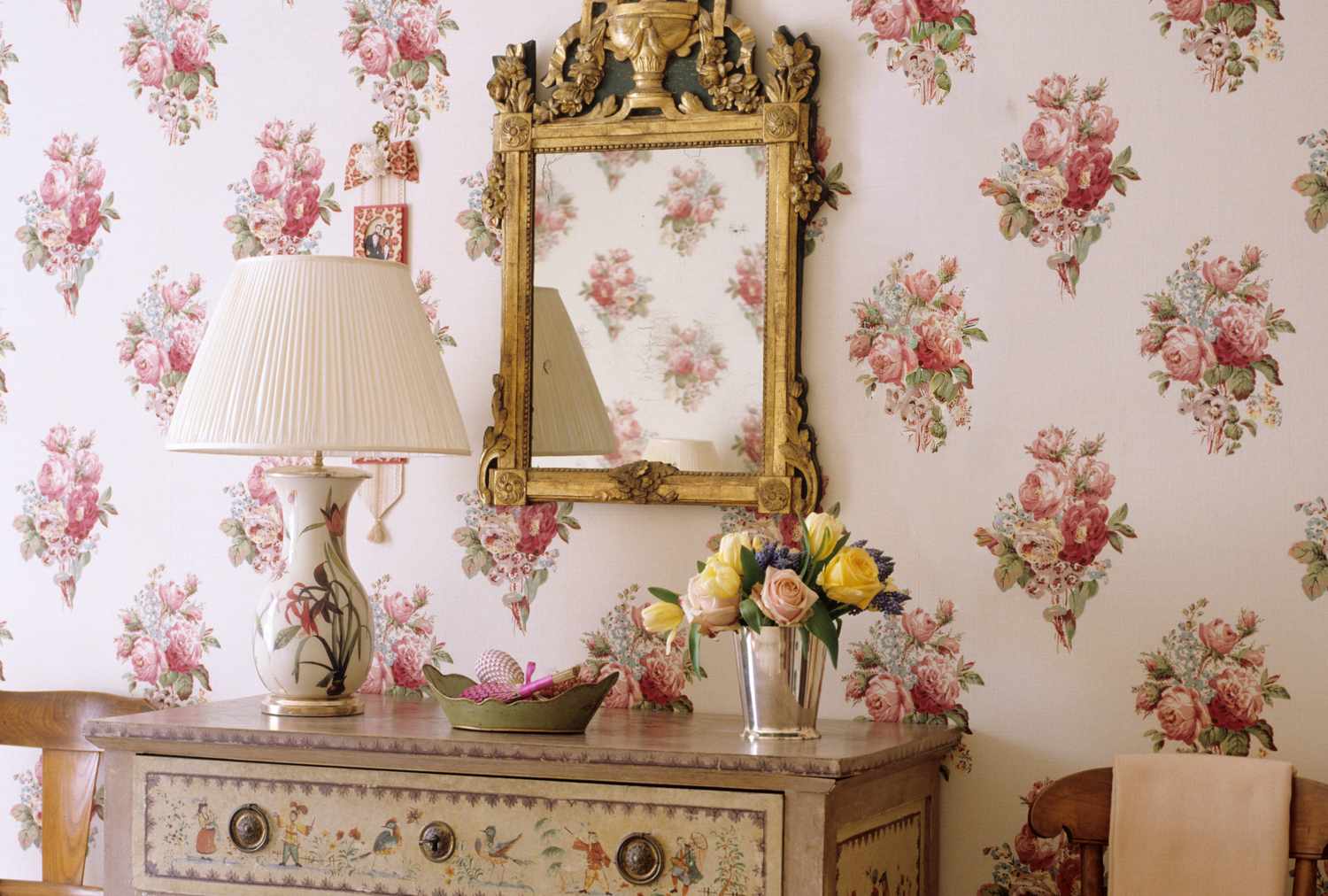 A vintagestyle dresser with a lamp vase of flowers and an ornate mirror on a floralpatterned wall
