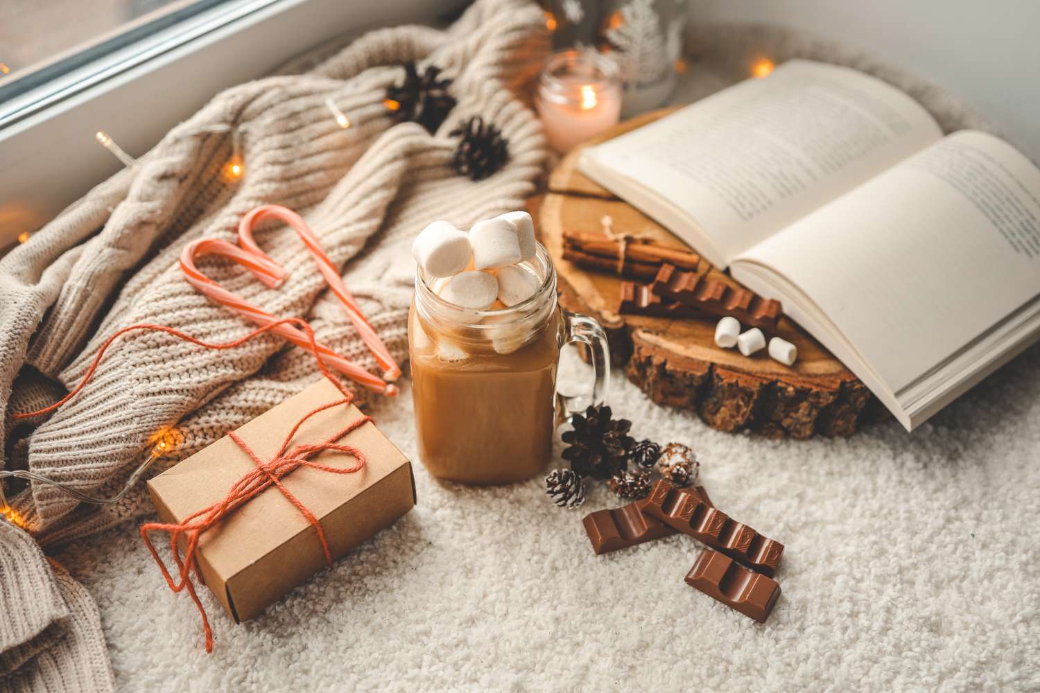 A cozy setup with a mug of hot cocoa topped with marshmallows an open book chocolate pieces and winterthemed decorations near a window