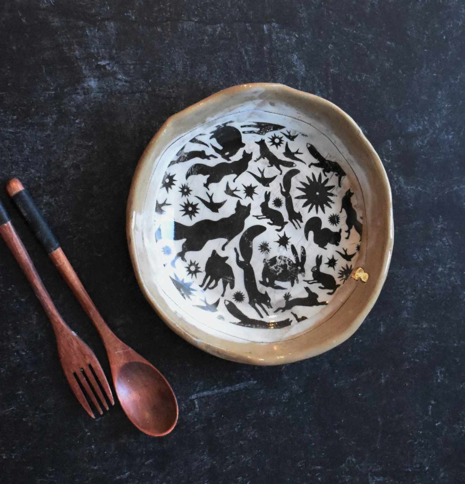A ceramic bowl with intricate black designs of animals and symbols next to a wooden fork and spoon
