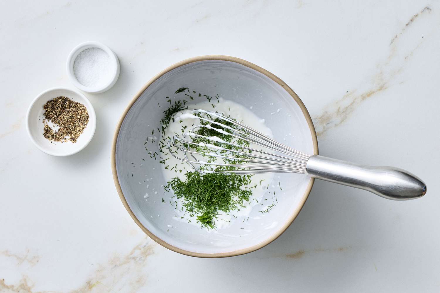 A bowl with sour cream, dill, and a whisk on a countertop, with small dishes of seasoning nearby