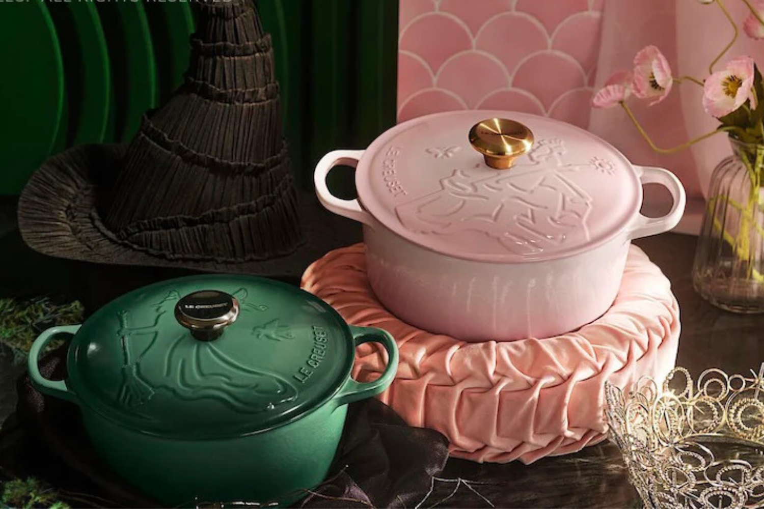 Two cast iron cookware pots one green and one pink displayed with decorative items including a witch hat and crown