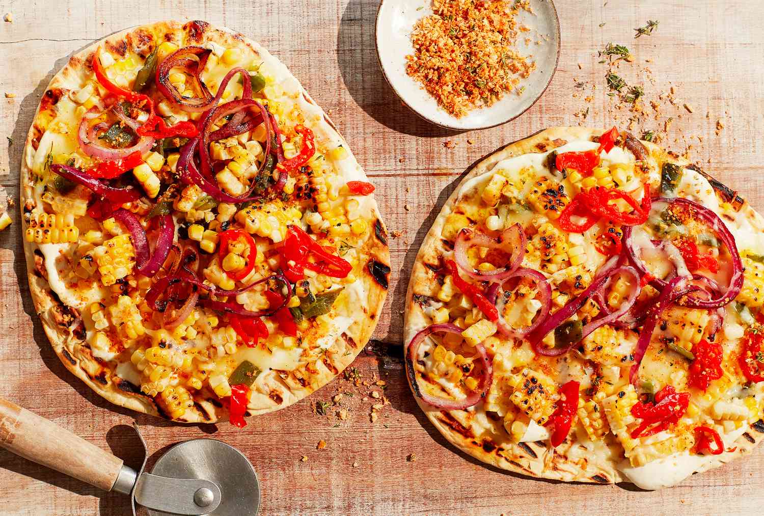 Two grilled pizzas topped with various vegetables and spices, with a pizza cutter and bowl of seasoning on a wooden surface