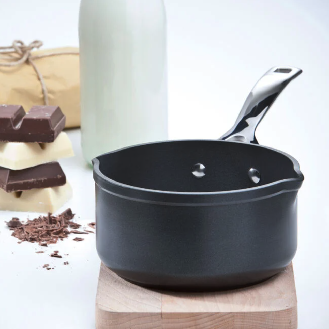 A kitchen saucepan on a wooden stand with chocolate, a milk bottle, and ingredients in the background