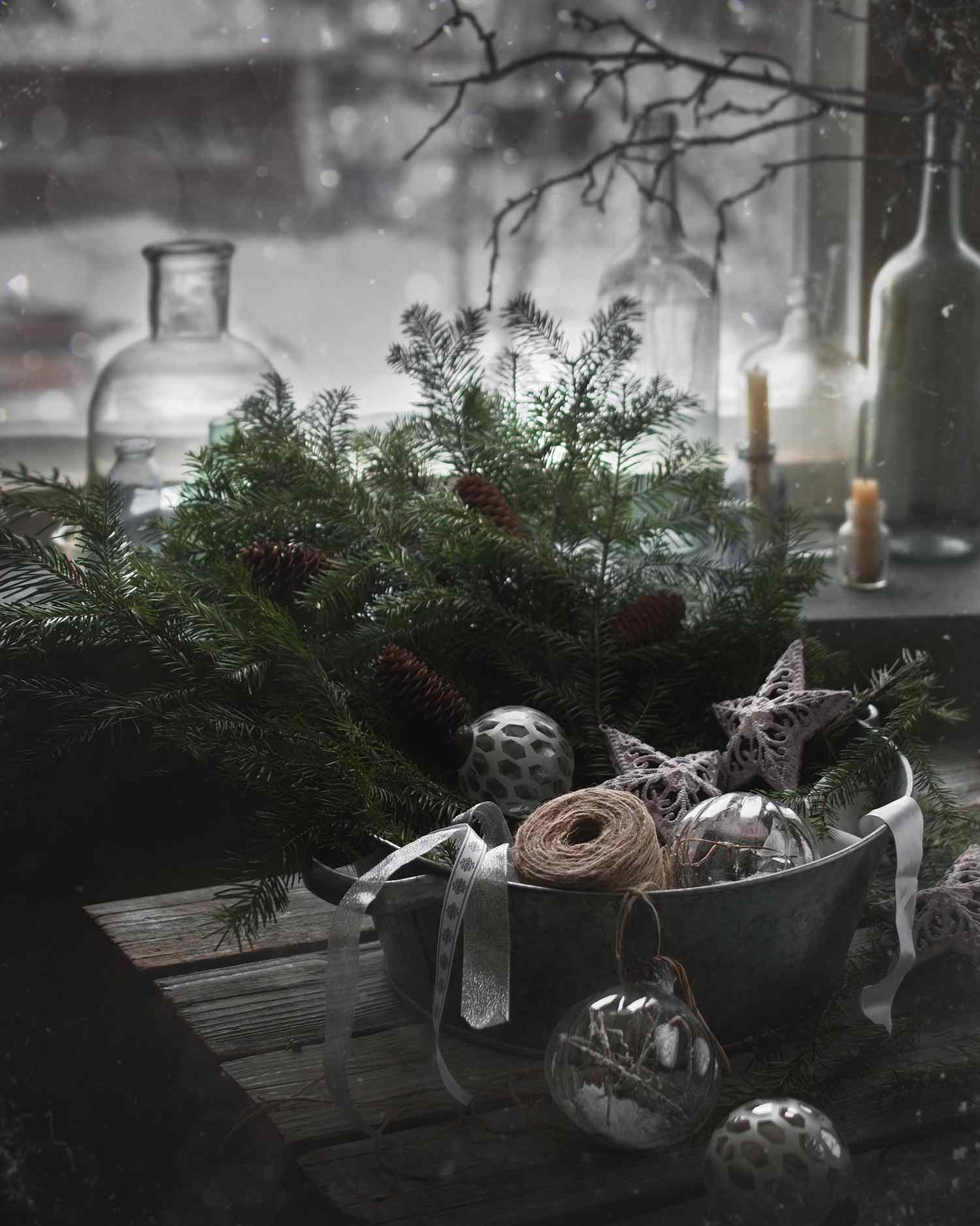 A rustic holidaythemed arrangement with greenery ornaments ribbon and candles set next to a window displaying a snowy scene