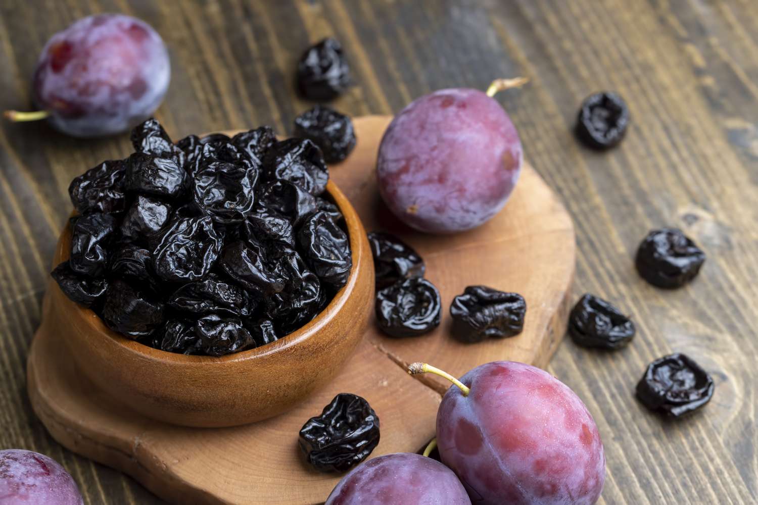 A wooden bowl containing dried prunes next to fresh plums on a wooden surface