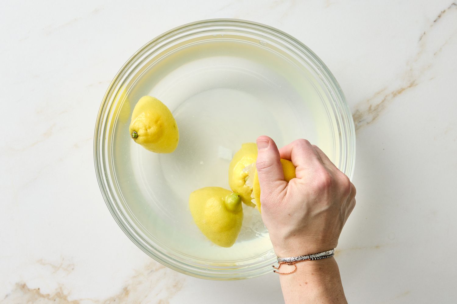 A hand holding a lemon submerged in a bowl of water containing other lemons