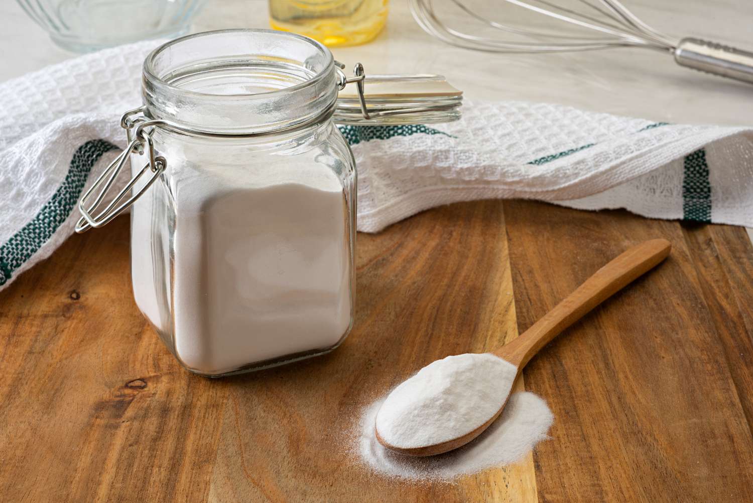 Jar of powdered ingredient and wooden spoon on a wooden surface with a towel and kitchen utensils in the background