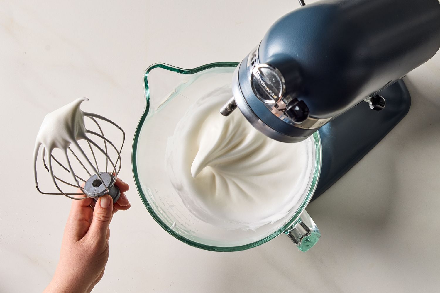 Whipped cream in a mixing bowl with a hand holding the whisk attachment